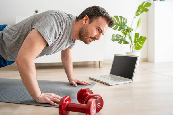 Ο άνθρωπος κάνει άσκηση, ενώ βλέποντας φροντιστήριο στο φορητό υπολογιστή στο σπίτι - Φωτογραφία, εικόνα