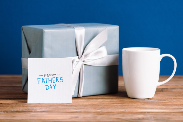 szelektív fókusz üdvözlő kártya felirattal boldog apák napja, fehér csésze és ajándék doboz fehér íj elszigetelt kék  - Fotó, kép