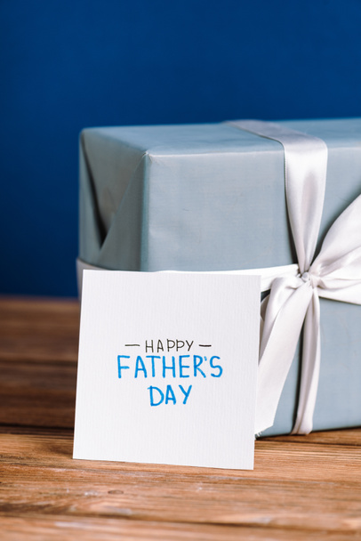 selective focus of greeting card with lettering happy fathers day and gift box with white bow on wooden table isolated on blue - Fotoğraf, Görsel