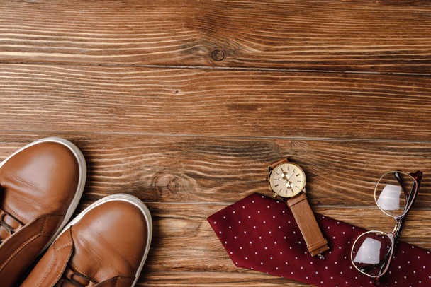 Top view of mens brown casual shoes, polka dots red tie, wristwatch and glasses on wooden background - 写真・画像