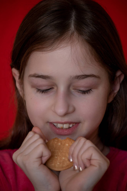 portrait of beautiful, lovely, l child girl, bites cookies on red background. Girl eating cookies. Girl 9 years old portrait. The concept of food and man. Teenager - Photo, Image