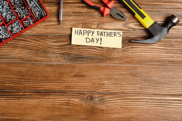 Pohled shora na blahopřání s ručně psaným nápisem Šťastný den otců, kladivo, kleště, šroubovák, matice a šrouby na dřevěném pozadí - Fotografie, Obrázek