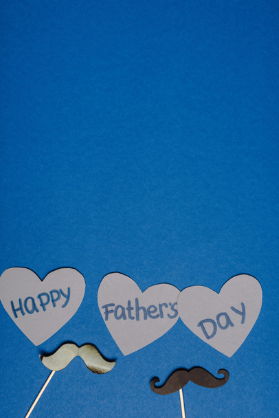 top view of decorative black mustache and paper cutted grey hearts with lettering happy fathers day on blue background - Foto, Imagem