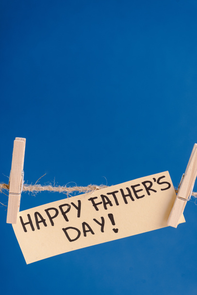 Tarjeta de felicitación beige con letras negras feliz día de los padres colgando de la cuerda con pinzas de ropa aisladas en azul
 - Foto, Imagen