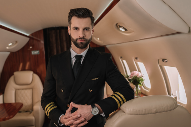 piloto guapo, seguro de sí mismo en uniforme mirando a la cámara mientras está de pie en jet privado
 - Foto, Imagen