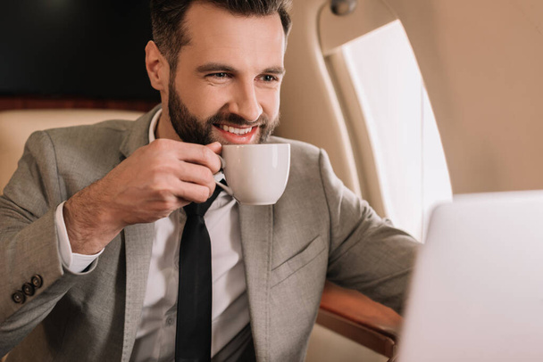 улыбающийся бизнесмен пьет кофе и смотрит на ноутбук в частном самолете
 - Фото, изображение