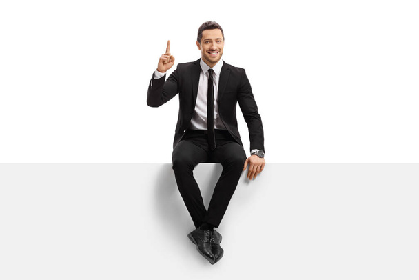 Full length portrait of a young man in a suit sitting on a panel and pointing upwards isolated on white background - Photo, Image