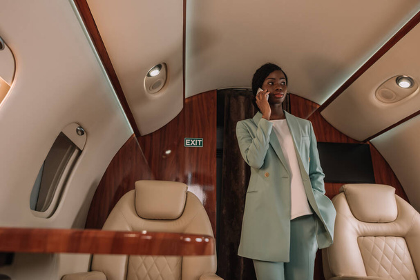 young, confident african american businesswoman talking on smartphone while standing in private jet - Foto, afbeelding