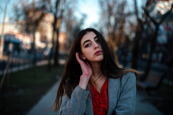 Girl in a red blouse and a gray cardigan on a blurry city background in early spring - Foto, Imagen
