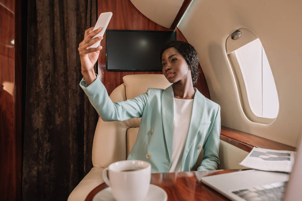 attractive african american businesswoman taking selfie on smartphone in private plane - Foto, afbeelding