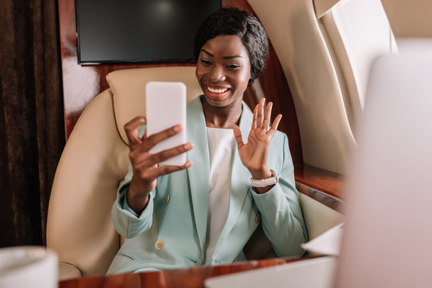 Selektiver Fokus einer glücklichen afrikanisch-amerikanischen Geschäftsfrau, die während eines Videogesprächs auf dem Smartphone winkt - Foto, Bild