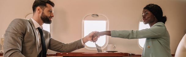 plano panorámico de atractiva mujer de negocios afroamericana estrechando la mano con hombre de negocios en avión
 - Foto, imagen