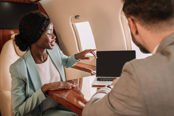 vue arrière de l'homme d'affaires près souriant afro-américaine femme d'affaires pointant avec le doigt et ordinateur portable avec écran blanc
 - Photo, image