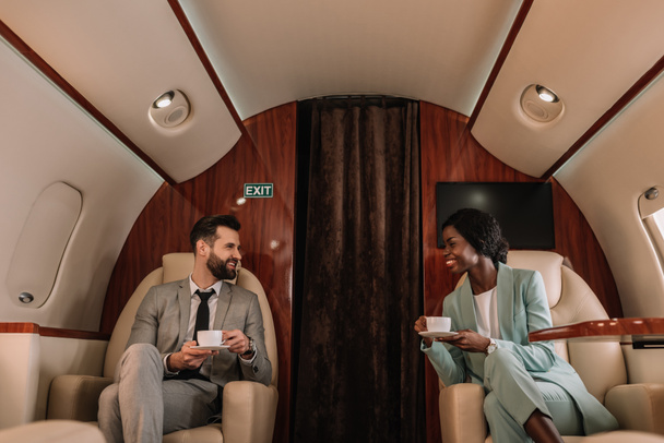 two smiling multicultural businesspeople talking while holding cups of coffee in plane - Photo, Image