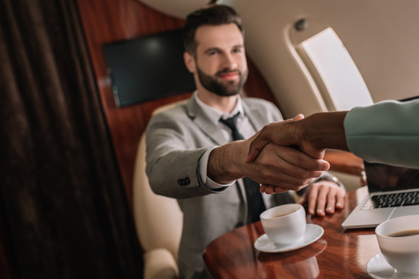 vista recortada de la mujer de negocios afroamericana estrechando la mano con hombre de negocios guapo en avión
 - Foto, imagen