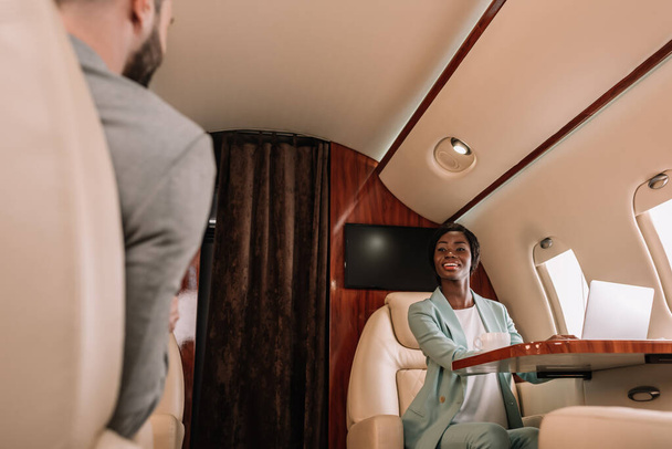zpět pohled na podnikatele při pohledu na usmívající se africké americké ženy při cestování v letadle - Fotografie, Obrázek