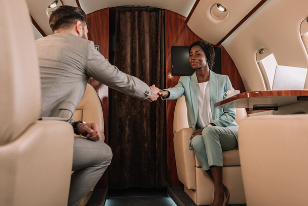 enfoque selectivo de dos empresarios multiculturales estrechando la mano mientras viajan en avión privado
 - Foto, Imagen