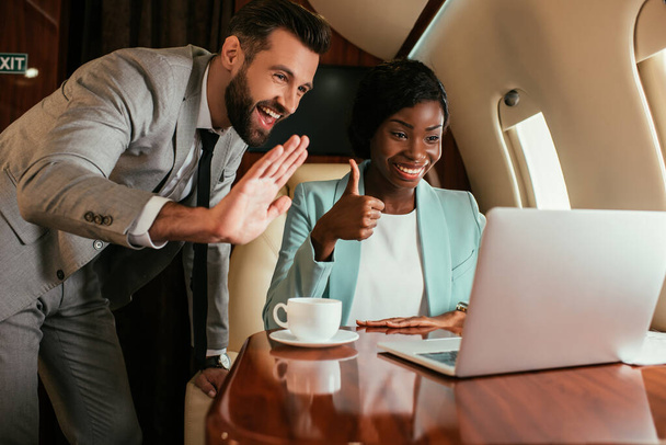 cheerful businessman waving hand and african american businesswoman showing thumb up during video call in private plane - Фото, изображение
