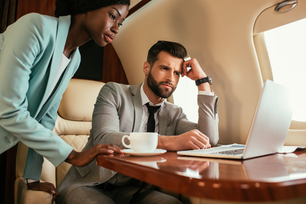 hombre de negocios serio y mujer de negocios afroamericana mirando portátil en jet privado
 - Foto, Imagen