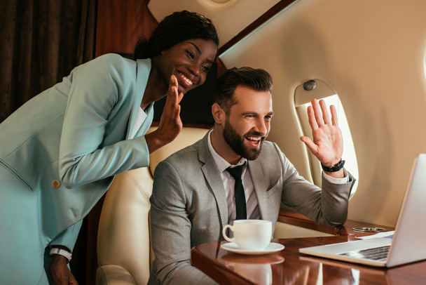 smiling muiticultural businesspeople waving hands while having video chat on laptop in private jet - Photo, Image