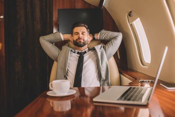 young businessman resting with hands behind head and closed eyes near laptop and cup of coffee in private plane - Foto, afbeelding