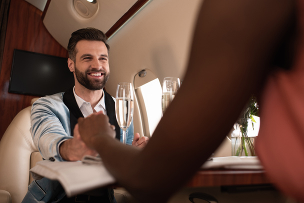 przycięty widok afrykańskiej amerykanki trzymającej się za ręce z uśmiechniętym eleganckim mężczyzną przy stole w prywatnym samolocie - Zdjęcie, obraz