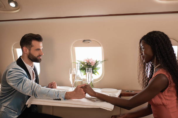 feliz, elegante pareja interracial tomados de la mano y mirándose unos a otros en la mesa servida en jet privado
 - Foto, Imagen