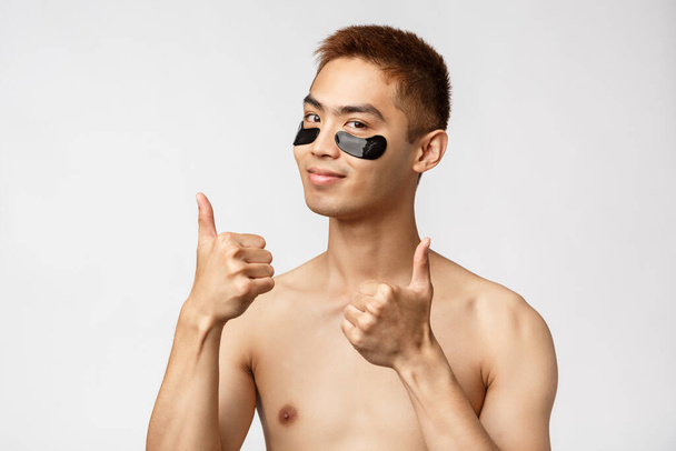 Beauty, people and lifestyle concept. Portrait of sassy good-looking asian man with naked torso, show thumbs-up in approval, use eye-patches for skincare routine, smiling satisfied, white background - 写真・画像
