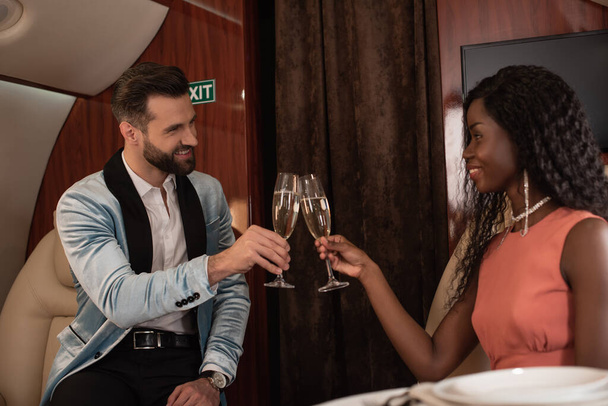 happy, elegant interracial couple clinking glasses of champagne in private plane - Фото, изображение