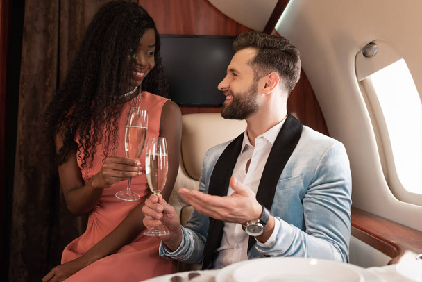 smiling interracial couple travelling in private jet and talking while holding glasses of champagne - Photo, Image