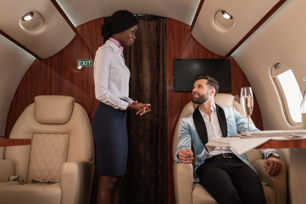 attraktive, lächelnde afrikanisch-amerikanische Stewardess, die neben einem gutaussehenden, eleganten Mann im Privatjet am gedeckten Tisch sitzt - Foto, Bild