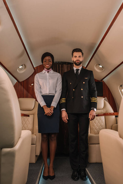 beau pilote confiant et souriant hôtesse afro-américaine regardant la caméra dans un avion privé
 - Photo, image