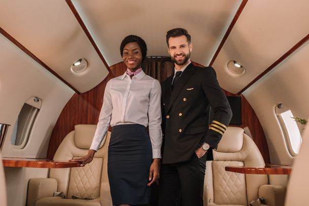 schöne, positive Pilotin, die mit der Hand in der Tasche neben lächelnder afrikanisch-amerikanischer Stewardess im Privatjet steht - Foto, Bild