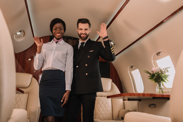 happy pilot and smiling afican american stewardess waving hands while looking at camera in private jet - Photo, Image