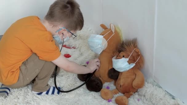 in a bright room, a boy with blue eyes in a medical mask, with a yellow t-shirt listens to brown toys of a horse and a hedgehog in a medium-sized stethoscope while sitting on the floor - Filmagem, Vídeo