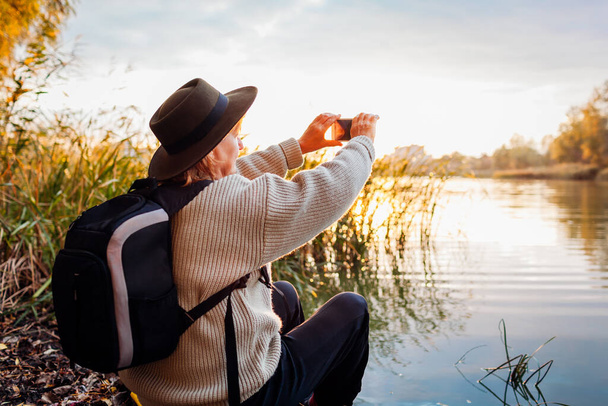 Tourist mit Rucksack, der Fotos mit dem Smartphone vom Fluss bei Sonnenuntergang macht. Frau reist bewundernd frühlingshafte Naturlandschaft. Die Welt erkunden - Foto, Bild