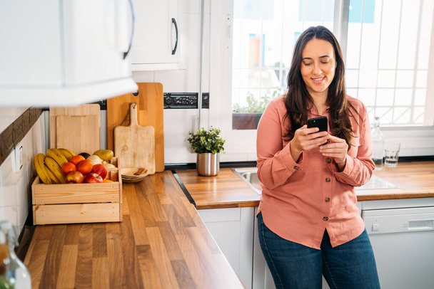 Jonge vrouw lacht typen sms-bericht op smartphone in haar keuken. - Foto, afbeelding