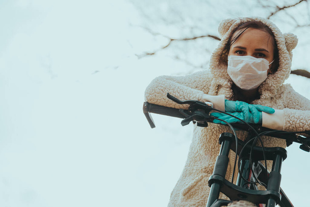 Portrait of young woman in medical mask and gloves with bicycle against sky. Adult female covering face to protect yourself from diseases on walk. Concept of threat of coronavirus epidemic infection. - Foto, Imagem