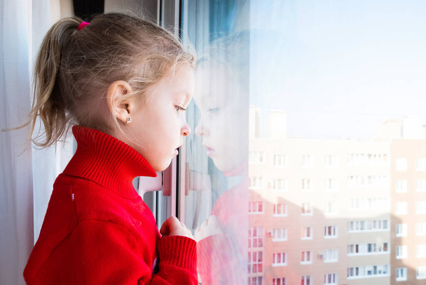 Très triste et grincheuse petite fille regarde par la fenêtre. Quarantaine pandémique. Reste à la maison. Verrouillage et enfants Covid-19
. - Photo, image