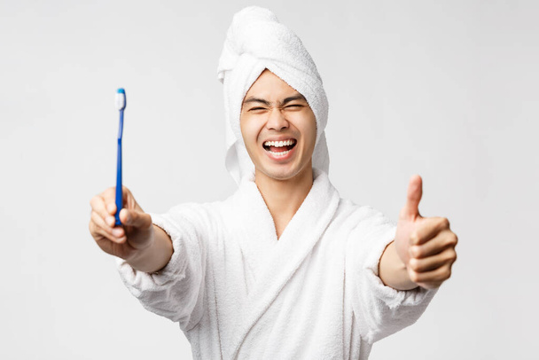 Conceito de beleza, spa e lazer. Retrato de entusiasta bonito asiático homem em roupão de banho e toalha de banho, mostrar escova de dentes com rosto satisfeito, fazer thumb-up em appoval, recomendo produtos de saúde
 - Foto, Imagem