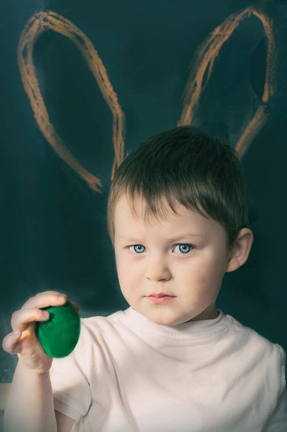 A very serious little boy is holding a green Easter egg in his hand. Rabbit ears are drawn in the background. Photo on a dark background, artistically processed. Vertical. - 写真・画像