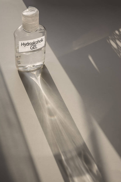 Une petite bouteille de gel hydro-alcoolique avec une étiquette et son ombre, sur une surface blanche. Rétroéclairage du soleil
 - Photo, image