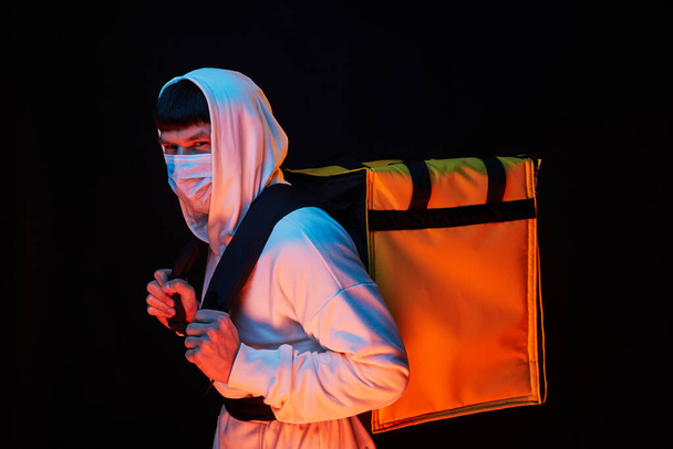 joven repartidor de ropa blanca con máscara de goma con una mochila amarilla sobre un fondo negro
 - Foto, imagen