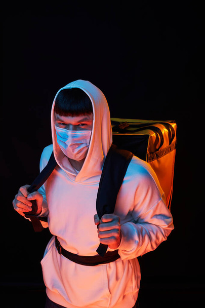 joven repartidor de ropa blanca con máscara de goma con una mochila amarilla sobre un fondo negro
 - Foto, Imagen
