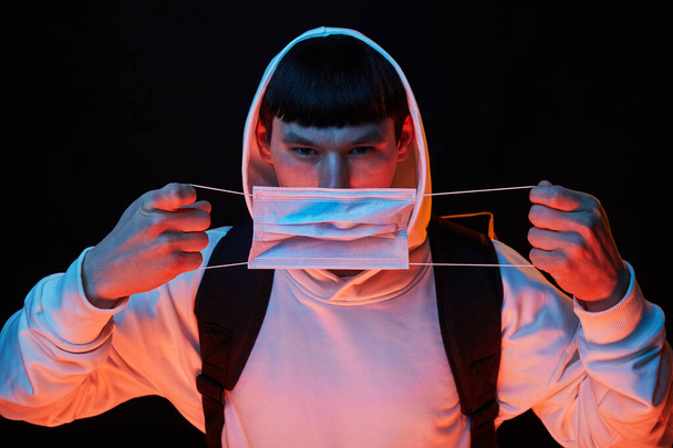 portrait young fashion guy in white hoodie in the hood robe in medicine mask in color lights on black background with backpack - Photo, Image