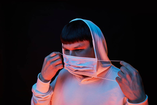 portrait young fashion guy in white hoodie in the hood robe in medicine mask in color lights on black background - Foto, Imagem