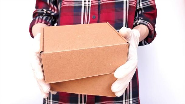 Courier in a checkered burgundy shirt with medical latex gloves safely delivers online purchases in brown cardboard boxes to the door on a white background during the coronavirus epidemic,COVID-19.The courier is holding brown cardboard boxes in his h - Photo, Image