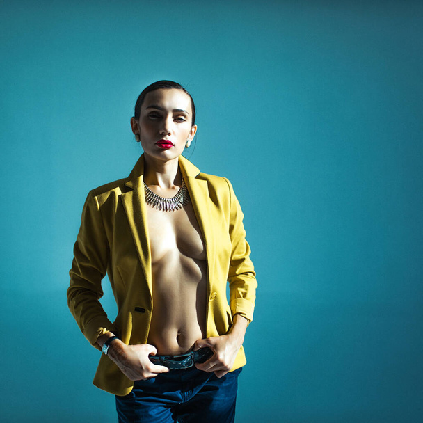 Young beautiful girl with dark hair neatly folded, well-dressed in a bright jacket on a naked body, pants and shoes, standing in the sunlight in an empty studio, blue background - Foto, Imagem