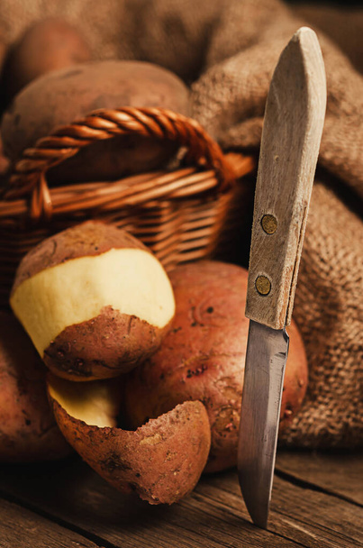 Cesta con patatas sobre una mesa de madera. El bodegón rural - la peladura con el cuchillo (la cocción) las patatas. comida tradicional ordinaria rústica
. - Foto, imagen