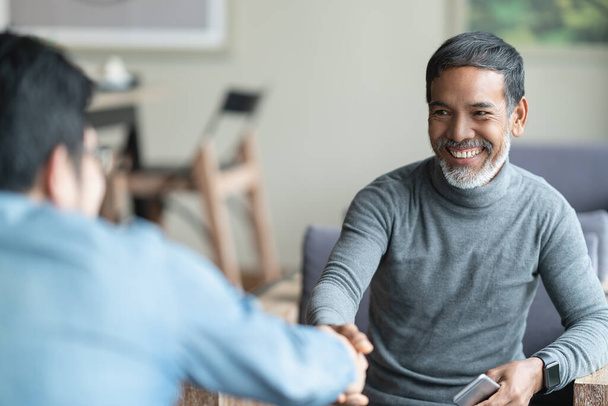 Confident mature Asian man sitting, smiling and shaking hand with partnership after making profitable agreement. Smart man handshake, deal and greeting concept. - Photo, Image